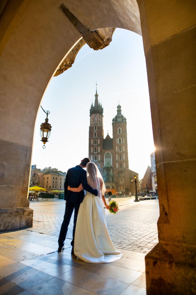 Para młoda obejmująca się na tle Bazyliki Mariackiej w Krakowie, widziana przez kamienny łuk z wiszącą latarnią, ciepłe światło słoneczne.