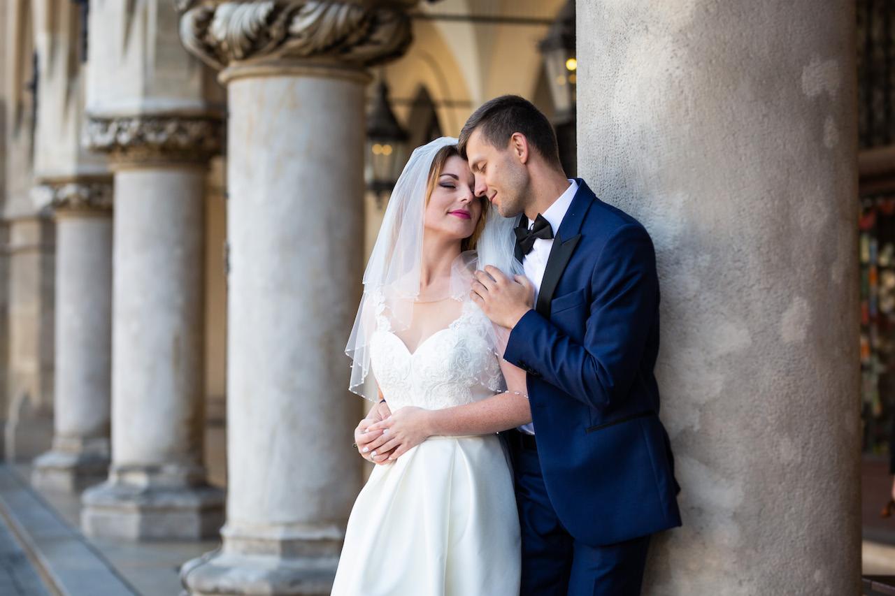 Panna młoda w białej sukni i welonie oraz pan młody w granatowym garniturze, stojący przy kolumnie i obejmujący się, z zamkniętymi oczami i uśmiechami na twarzach, na tle rzędu kolumn.