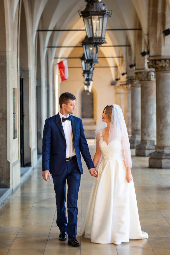 Para młoda, on w granatowym garniturze z muchą, ona w białej sukni ślubnej z welonem, spacerują trzymając się za ręce pod arkadami z kolumnami, w tle widoczna polska flaga.