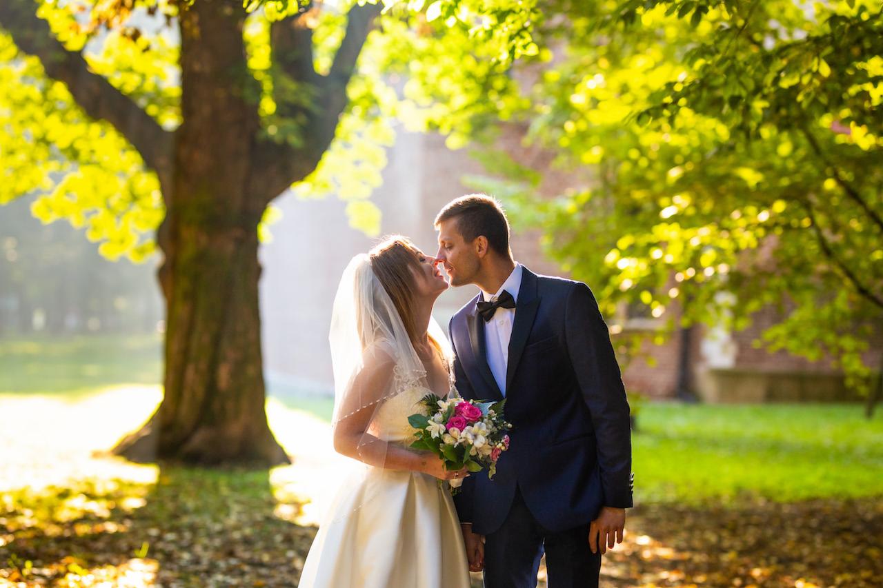 Para młoda w plenerze, pocałunek na tle drzew z zielonymi liśćmi i ceglanego budynku w oddali, panna młoda trzyma bukiet z różowych i białych kwiatów.