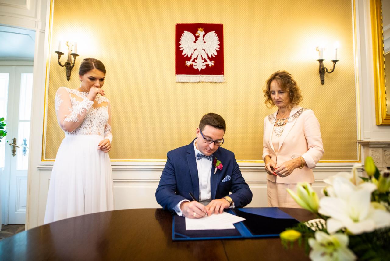 Para młoda podczas składania podpisu w Urzędzie Stanu Cywilnego, w tle godło Polski i urzędniczka.