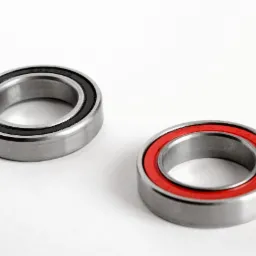 Dwa łożyska kulkowe, jedno z czarnym uszczelnieniem, drugie z czerwonym, leżące na białym tle, widoczne ślady smaru.