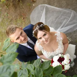 Para młoda, panna młoda w koronkowej sukni z bukietem czerwono-białych kwiatów, pan młody w granatowym garniturze, ujęcie z góry, widok przez liście krzewu.