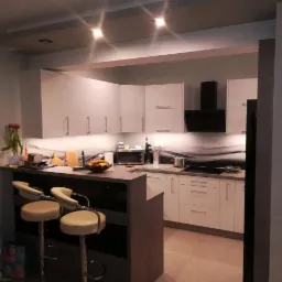 Jasna kuchnia z barkiem i hokerami, oświetlenie punktowe, dekoracyjny panel nad blatem, widok z perspektywy wejścia.