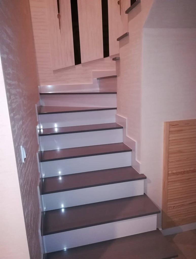 Nowoczesne schody wewnętrzne z szarymi stopniami i białymi podstopnicami, oświetlone punktowo, z dekoracyjną ścianą w tle.