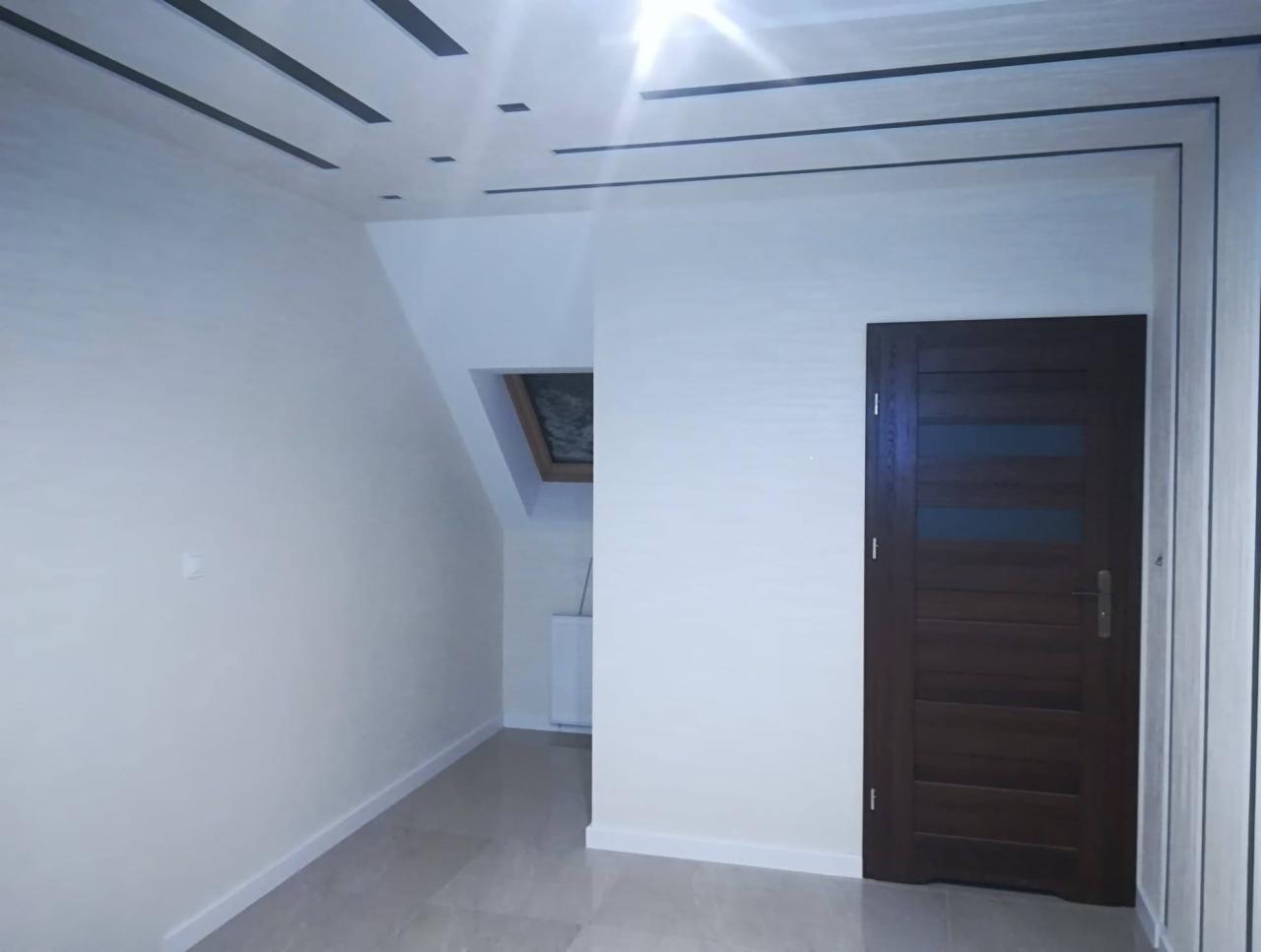 Wykończone wnętrze pokoju na poddaszu z drewnianymi drzwiami z szybą, szarą podłogą i białymi ścianami z czarnymi akcentami na suficie.