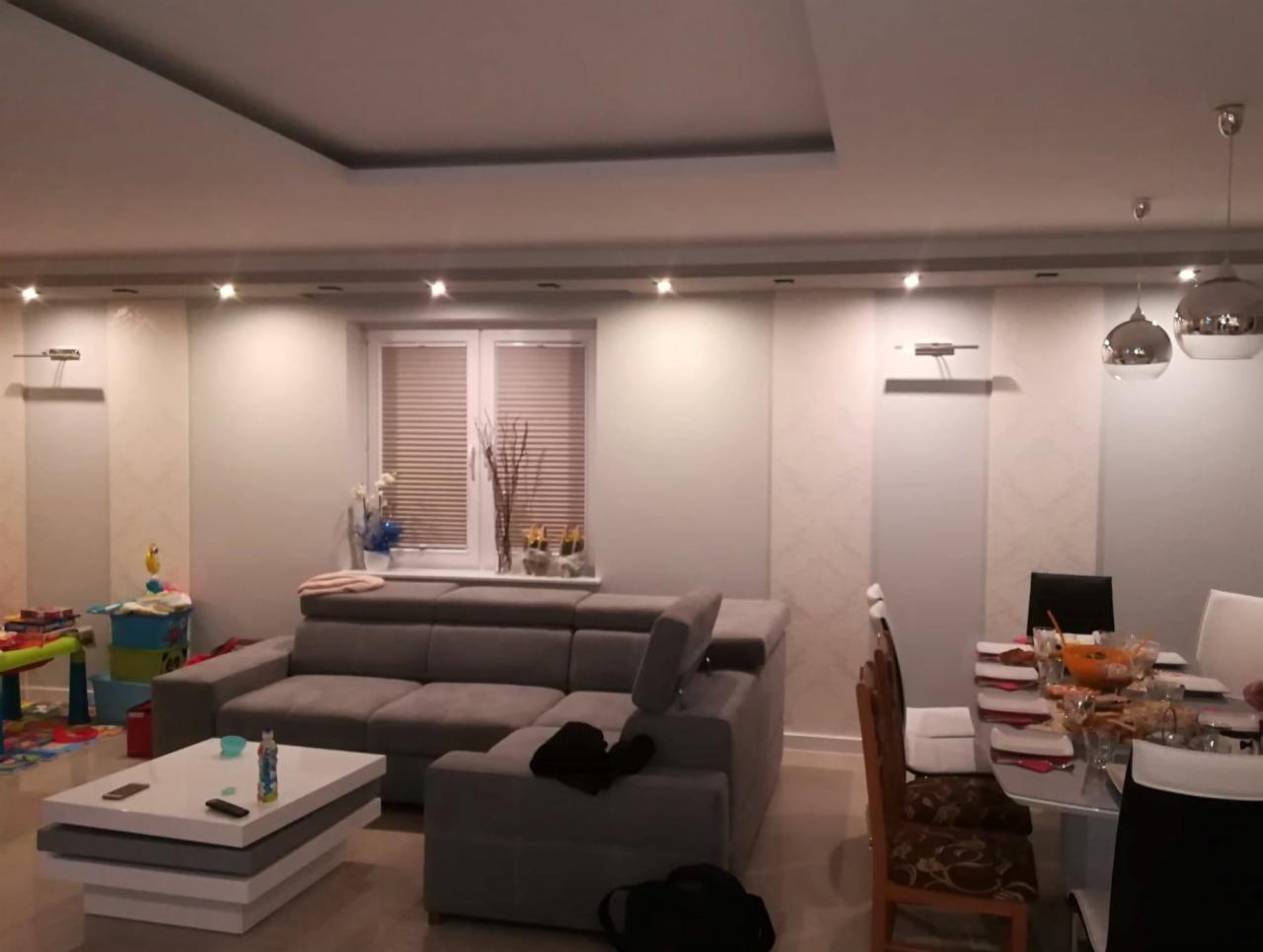 Jasne wnętrze salonu z szarą narożną sofą, białym stolikiem kawowym i stołem jadalnym z nakryciem, oświetlone punktowymi lampami sufitowymi i wiszącymi srebrnymi kulami, z widocznymi elementami...