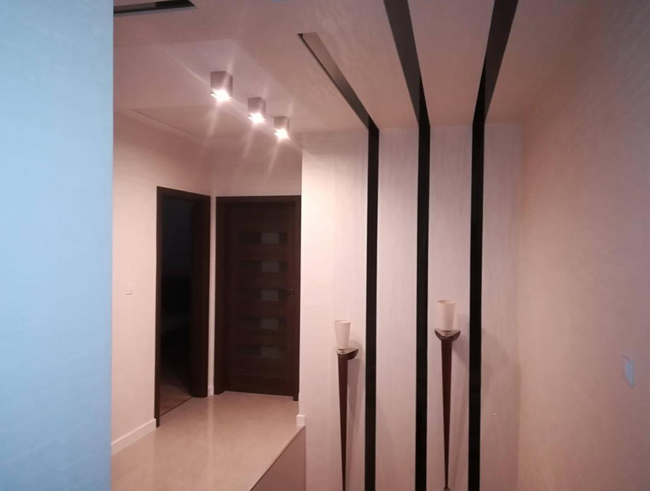 Korytarz z dekoracyjnymi czarnymi pasami na suficie i ścianach, oświetlenie punktowe sufitowe i wysokie świeczniki na ścianie, widok na otwarte drzwi do dalszej części mieszkania.