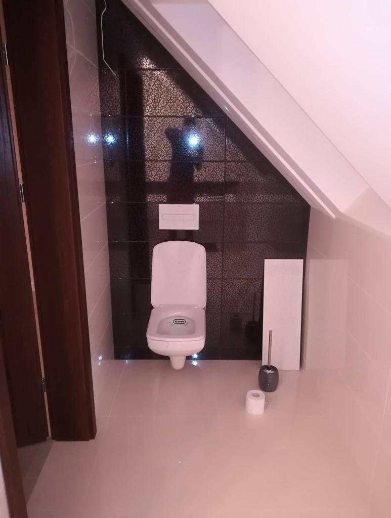 Nowoczesna łazienka na poddaszu z czarnymi płytkami mozaikowymi na ścianie, wiszącą toaletą z deską sedesową i akcesoriami w kolorze szarym.