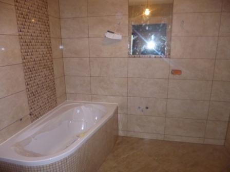 Jasna łazienka po remoncie z wanną obudowaną mozaiką, fragment ściany z dekoracyjną wstawką z płytek i lustro z widocznymi plamami.