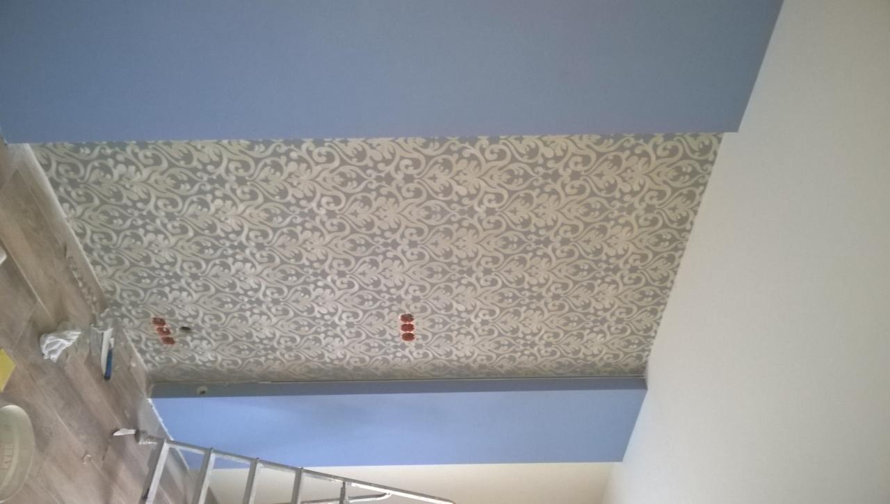 Dekoracyjny sufit w jasnych odcieniach szarości z wzorem roślinnym, obok niebieski pas i biała ściana, widoczne gniazdka elektryczne.