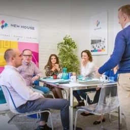 Zespół w biurze agencji nieruchomości podczas spotkania przy białym stole, w tle banery reklamowe z logo firmy New House oraz roślina doniczkowa.