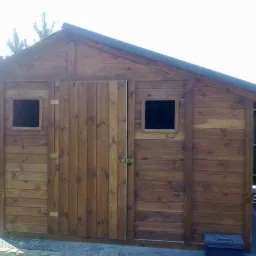 Gotowa, drewniana szopka narzędziowa z dachem dwuspadowym, małym oknem i drzwiami na zawiasach, umieszczona w ogrodzie.