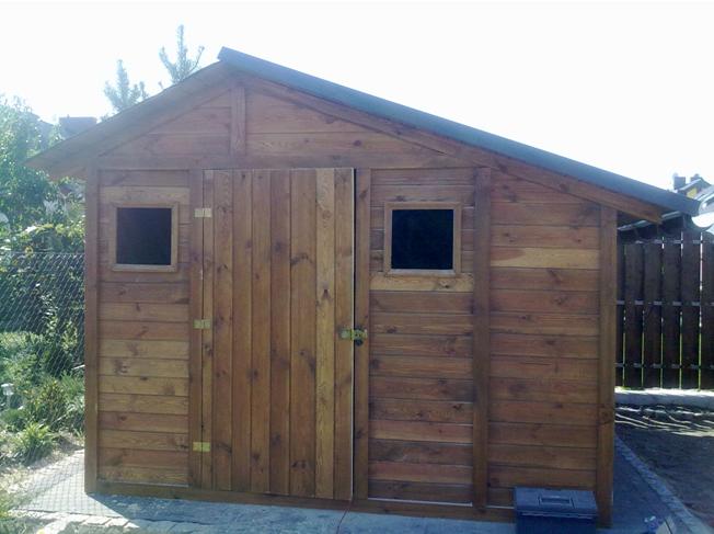 Gotowa, drewniana szopka narzędziowa z dachem dwuspadowym, małym oknem i drzwiami na zawiasach, umieszczona w ogrodzie.