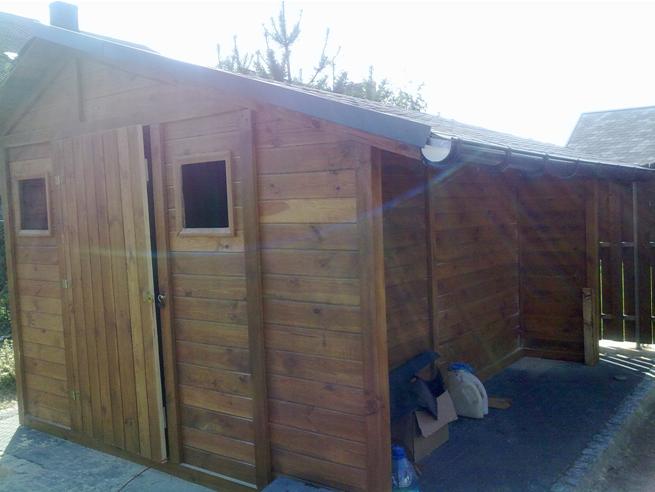 Drewniana szopka z przesuwnymi drzwiami i dwoma małymi okienkami, zadaszona z rynnami, widok z boku na tle ogrodu.