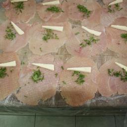 ASK sp z o.o. - Przygotowane plastry surowego mięsa, posypane ziołami i z kawałkiem sera, ułożone na drewnianej desce zabezpieczonej folią spożywczą.