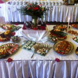 ASK sp z o.o. - Stół bankietowy z bogato zastawionym cateringiem: dekoracyjne kompozycje z owoców, warzyw, wędlin i serów, bukiet czerwonych kwiatów w szklanym wazonie, kieliszki z czerwonymi owocami, zapalona...