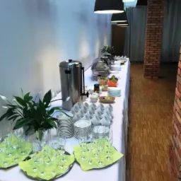 Stół cateringowy z podgrzewaczami, szklankami na zielonych serwetkach, stosami talerzy i kwiatami w doniczce, przygotowany do obsługi gości podczas wydarzenia firmowego.