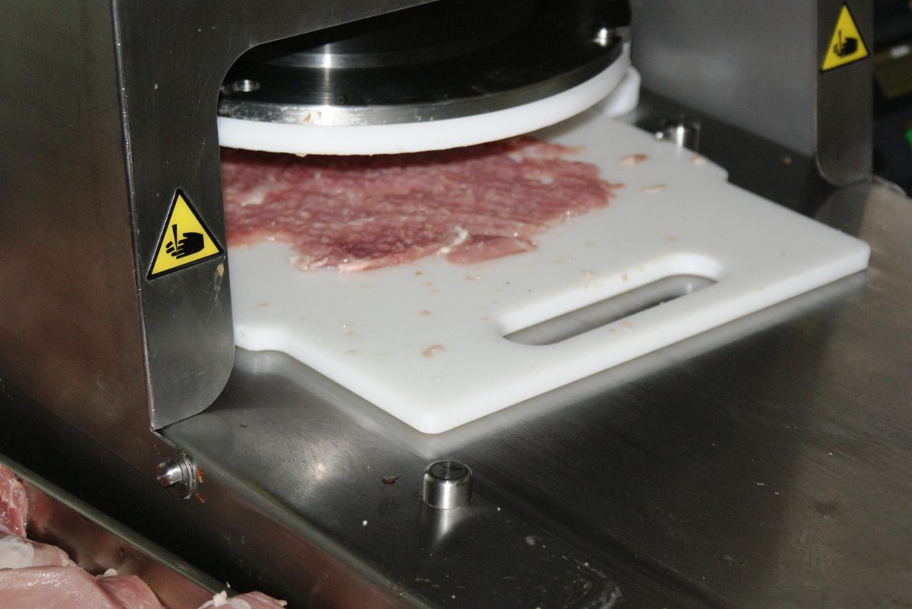 Automatyczna krajalnica do wędlin tnąca salami na cienkie plasterki na białej desce, widoczne oznaczenia ostrzegawcze na obudowie urządzenia.