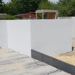 Świeżo otynkowane, białe ściany w trakcie budowy domu, widoczne z zewnątrz, z elementami instalacji wystającymi ze ściany, na pierwszym planie szara kostka brukowa, w tle palety i gruz.