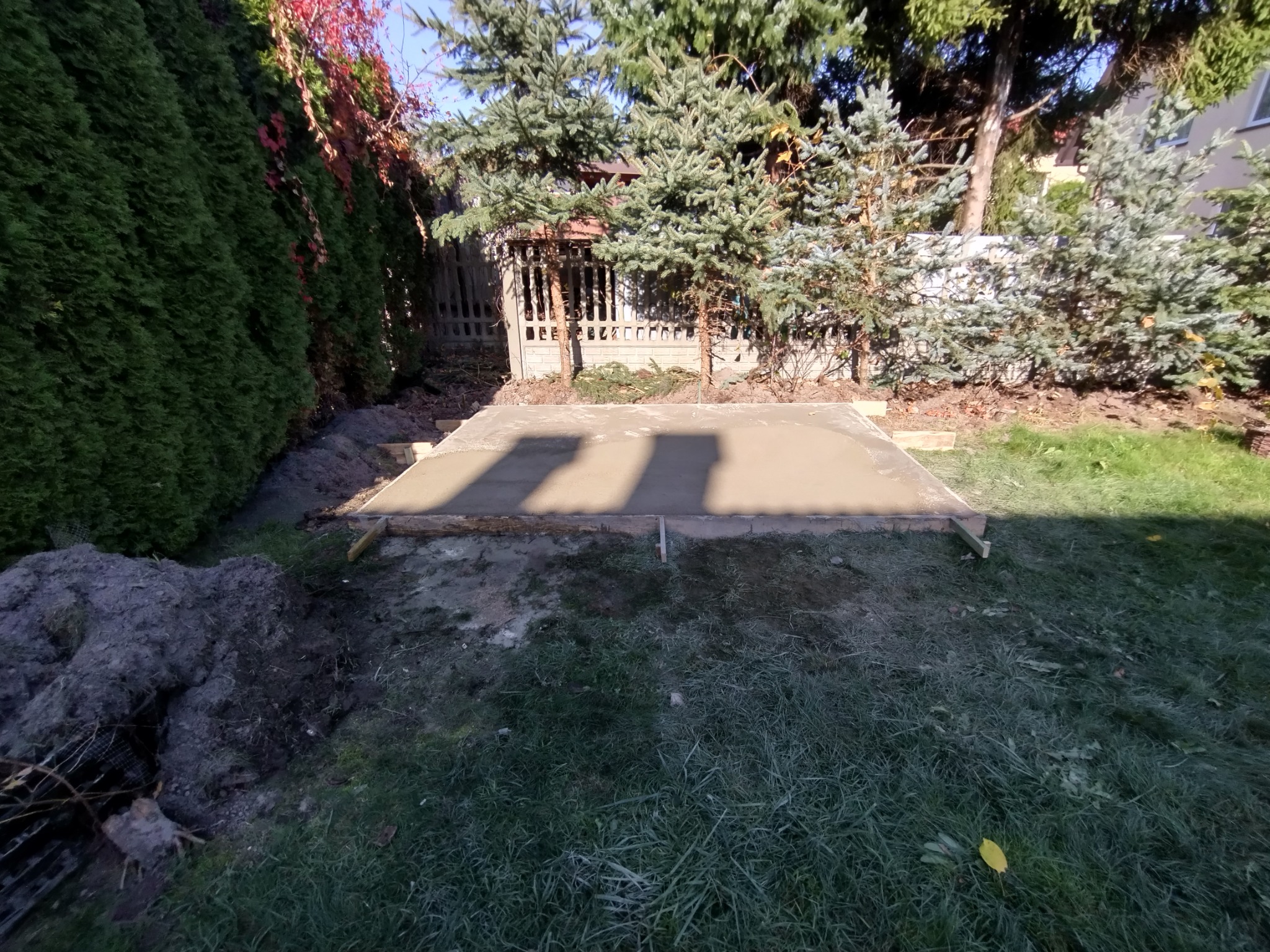 Świeżo wylana, jeszcze mokra betonowa płyta fundamentowa w drewnianym szalunku na trawiastym terenie działki, z widoczną kupą ziemi z boku i zielonymi drzewami w tle.