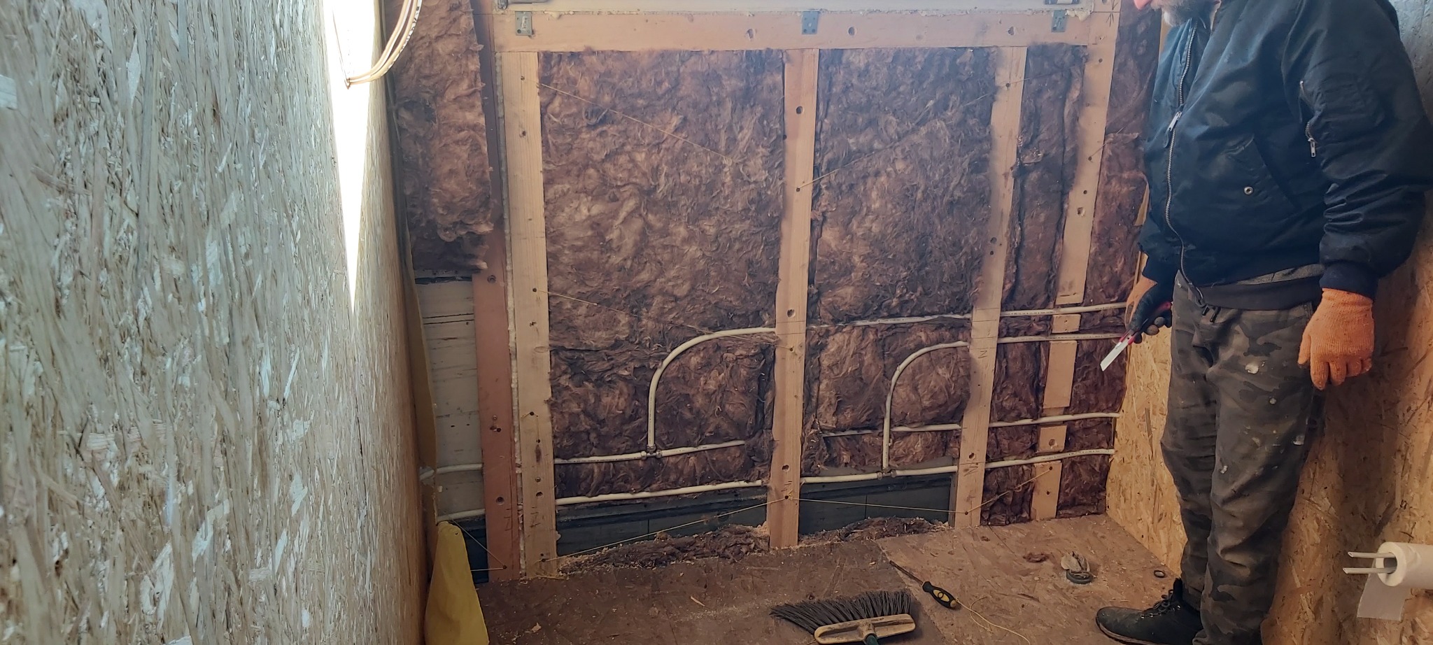 Wnętrze domu w trakcie ocieplania wełną mineralną, widoczne drewniane belki konstrukcyjne, instalacja elektryczna, płyta OSB na ścianie oraz mężczyzna w roboczym ubraniu trzymający nóż do cięcia...