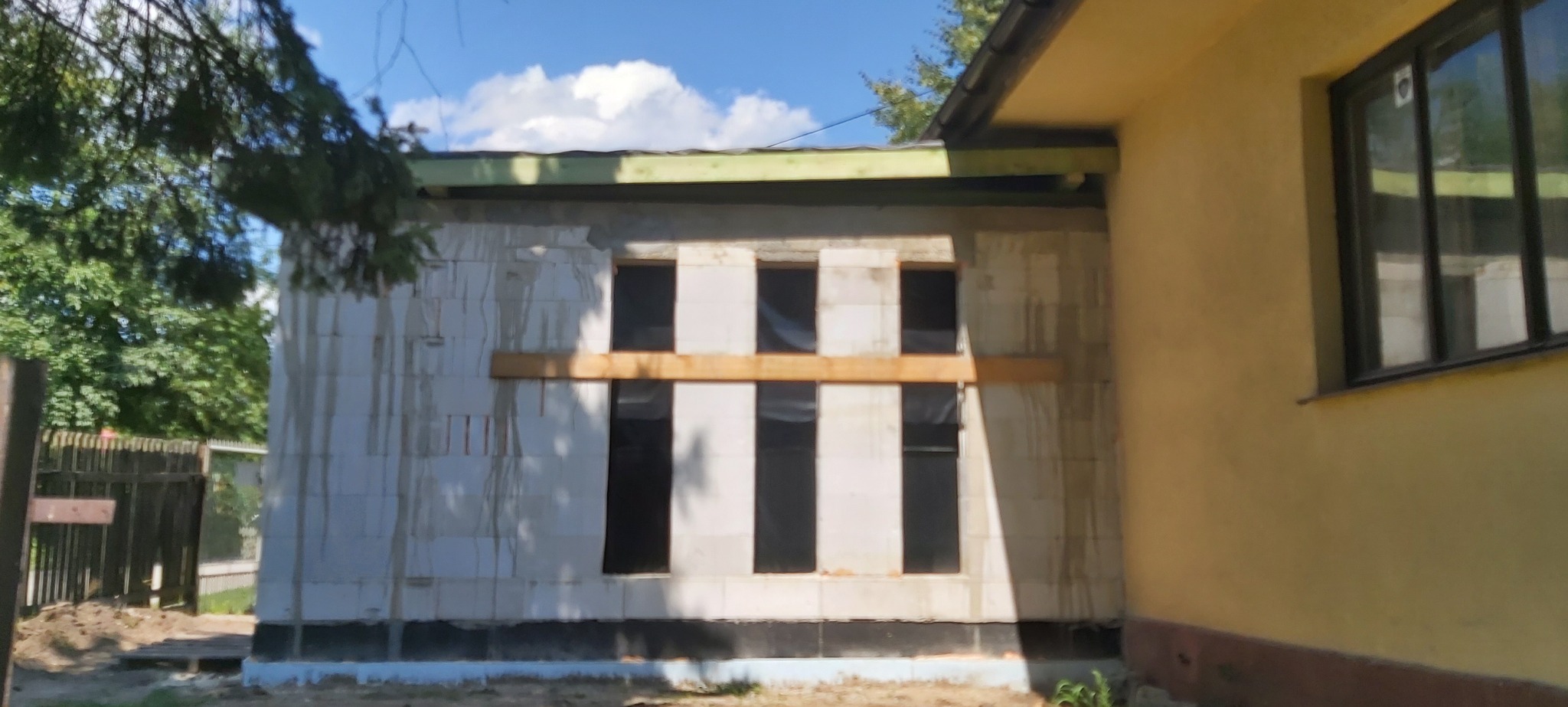 Wznoszenie ściany z bloczków na budowie domu, widoczne otwory okienne zabezpieczone folią, drewniana belka podtrzymująca nadproże, fragment istniejącego budynku z oknem w tle, drzewa i błękitne niebo.