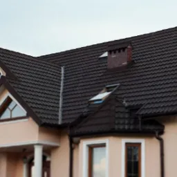 Dom jednorodzinny z brązowym dachem z blachy, widoczne rynny, okna dachowe, komin z cegły i balkon z ozdobną balustradą.