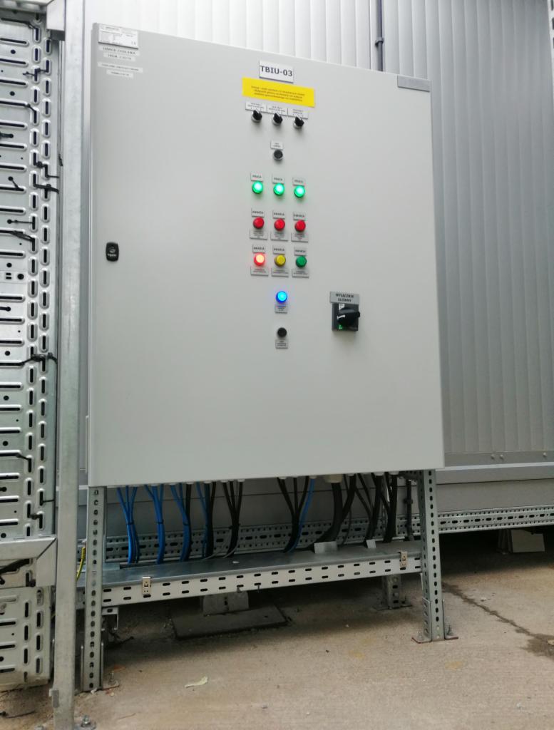 Szara szafa sterownicza TBIU-03 z włącznikiem, kontrolkami LED (zielone, czerwone, żółte, niebieskie) i podłączonymi przewodami, umieszczona na metalowej konstrukcji w pomieszczeniu technicznym.