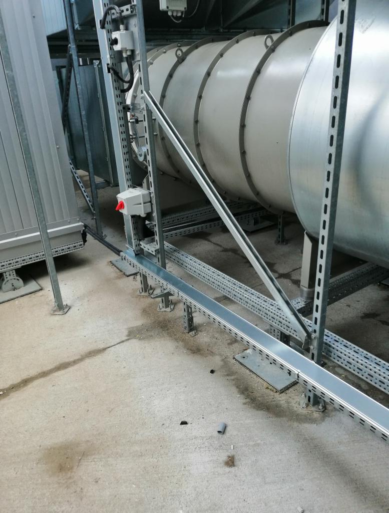 Szara, metalowa rura wentylacyjna z pierścieniami wzmacniającymi, wsparta na metalowej konstrukcji z perforowanymi profilami, widoczna instalacja elektryczna z puszką z czerwonym przyciskiem.