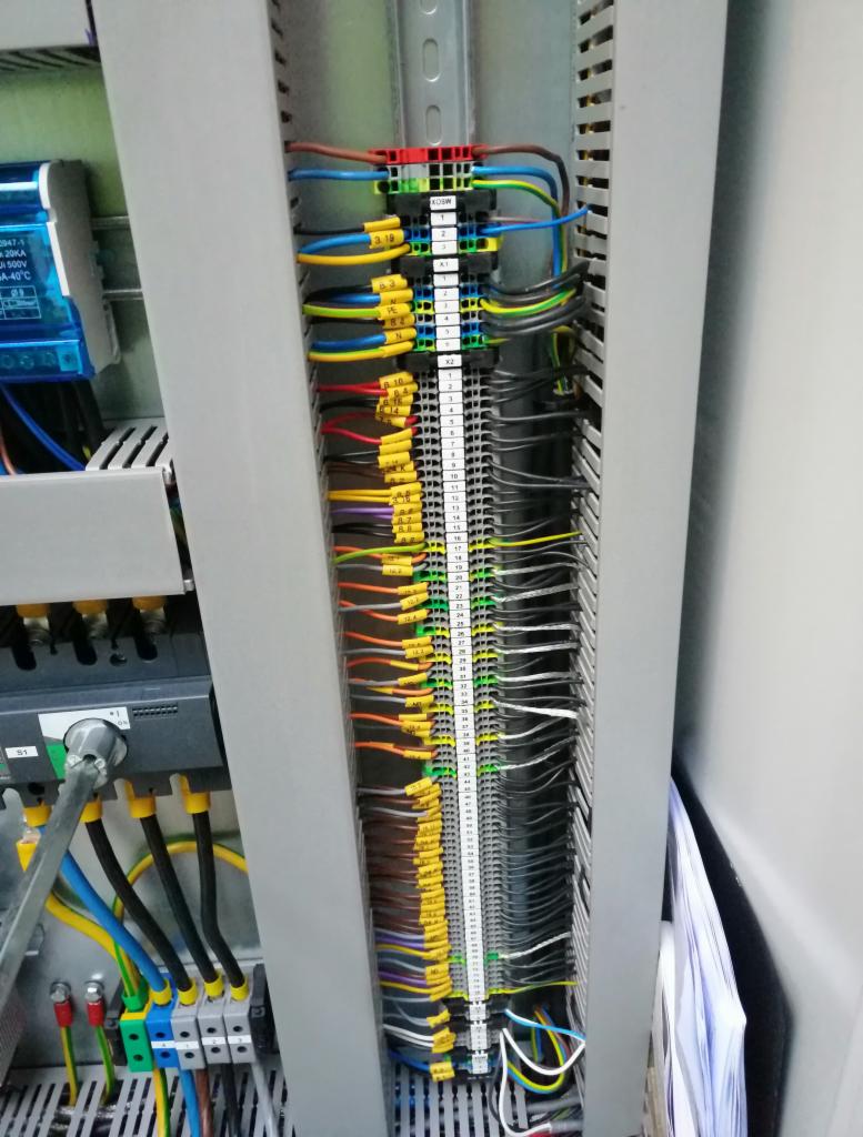 Szczegółowe ujęcie szafy sterowniczej z licznymi przewodami oznaczonymi żółtymi etykietami, podłączonymi do listew zaciskowych. Widoczne również elementy montażowe i okablowanie w kolorach: czarnym...