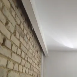 Ściana z cegły rozbiórkowej połączona z gładką, pomalowaną na biało ścianą i sufitem, widoczna listwa sufitowa.