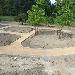 Progress Gliwice - Park w trakcie budowy: ścieżki z jasnego żwiru obramowane szarą kostką, przygotowane rabaty. Widoczne drzewa z podporami i teren z czerwonymi znacznikami.