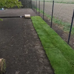 Układanie trawy z rolki wzdłuż ogrodzenia z siatki, widoczny przygotowany teren i zwinięta rolka trawy.