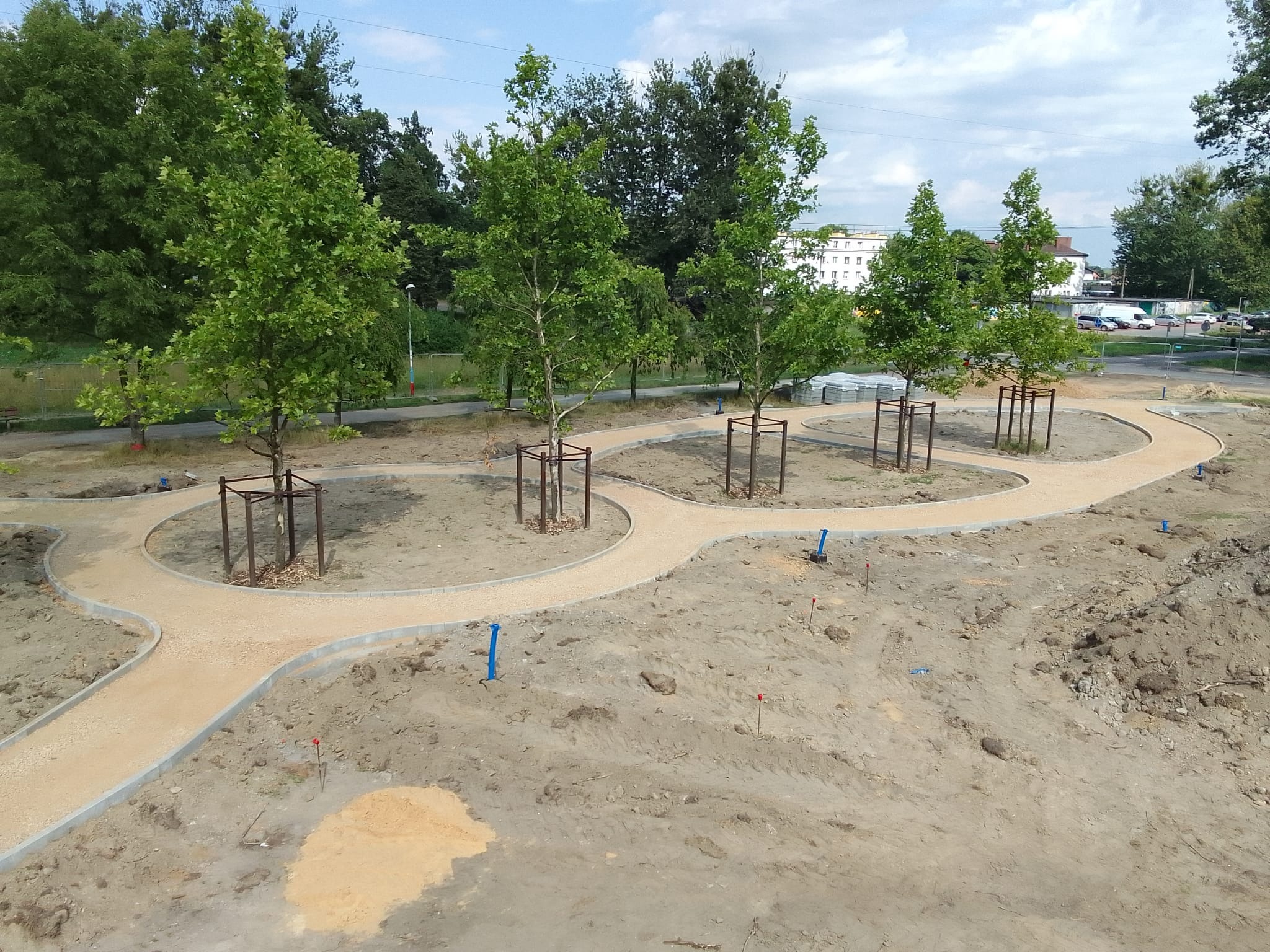 Parkowa ścieżka z jasnego żwiru, obrzeża z kostki, młode drzewa z zabezpieczeniami, teren przygotowany pod nasadzenia. Słoneczny dzień.