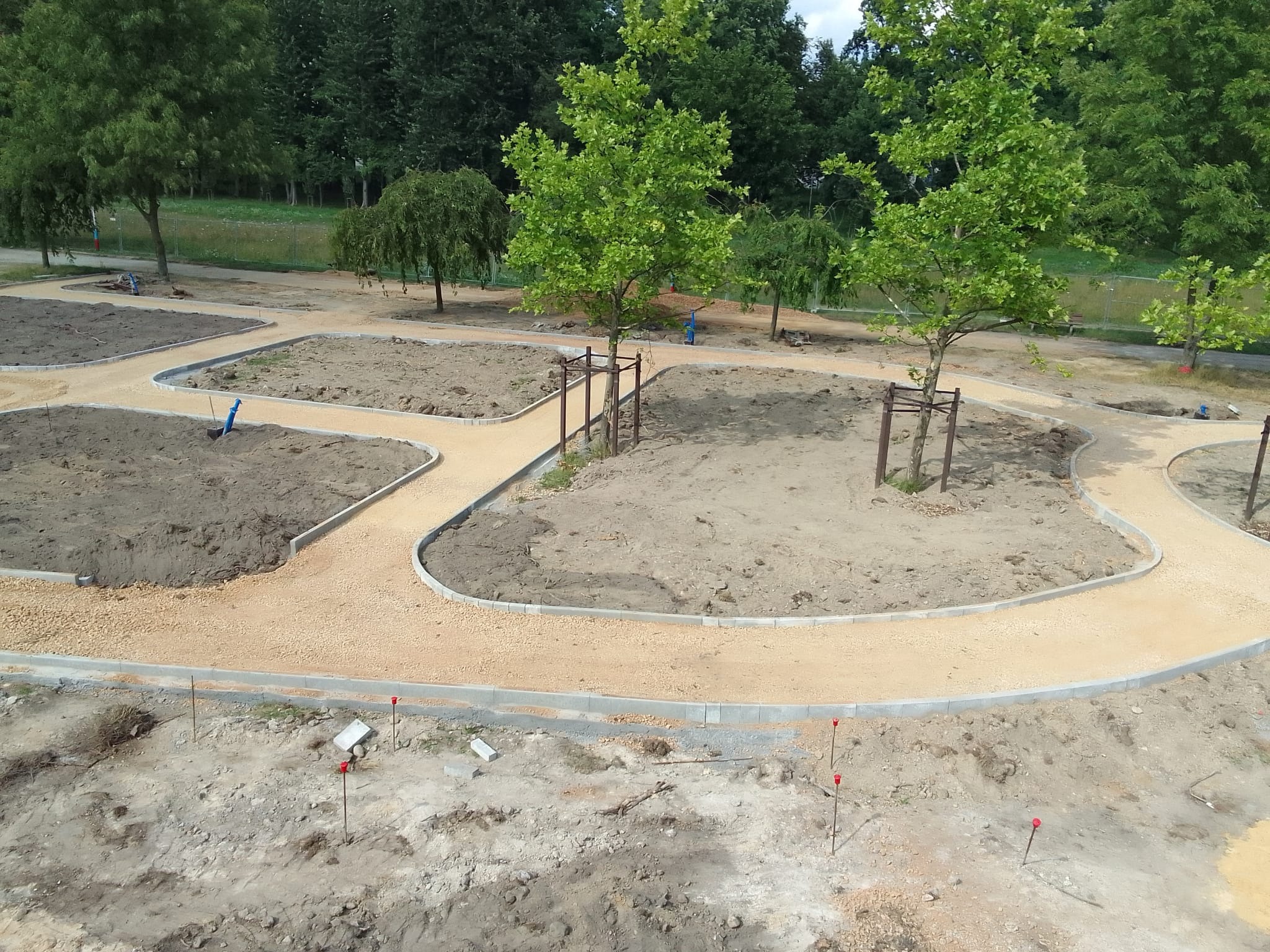 Park w trakcie budowy: ścieżki z jasnego żwiru obramowane szarą kostką, przygotowane rabaty. Widoczne drzewa z podporami i teren z czerwonymi znacznikami.