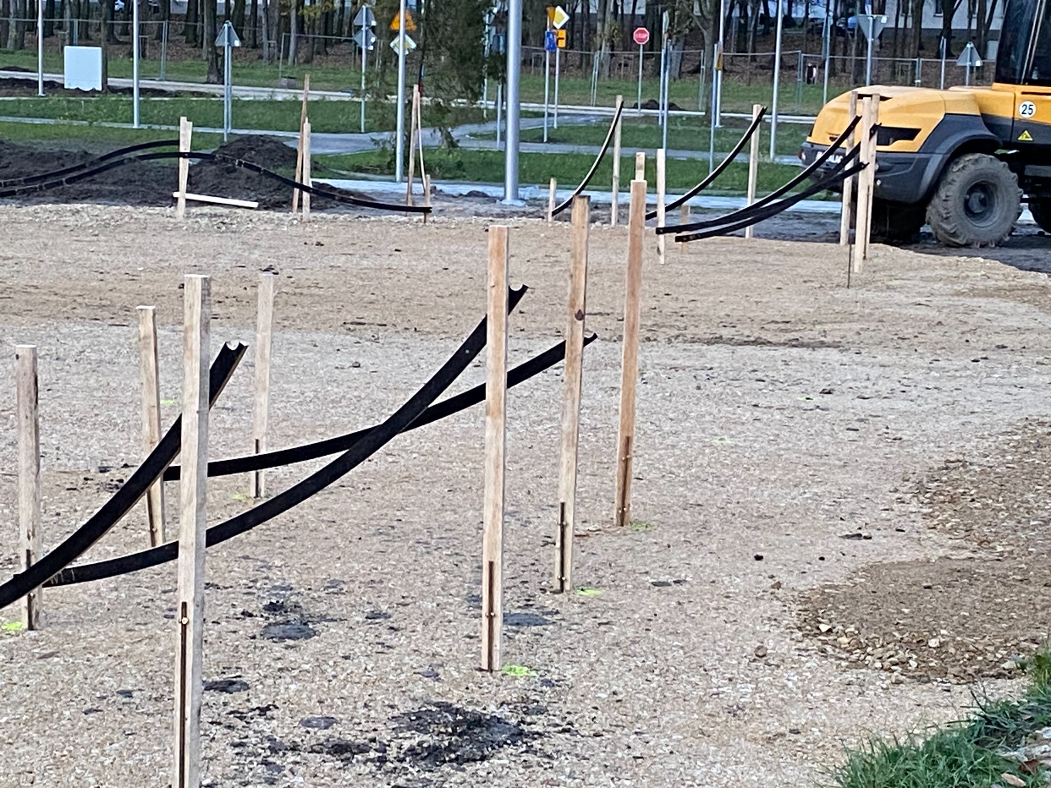 Przygotowanie terenu pod instalacje: drewniane paliki wyznaczają trasę czarnych rur, w tle widoczna maszyna budowlana i elementy infrastruktury miejskiej.