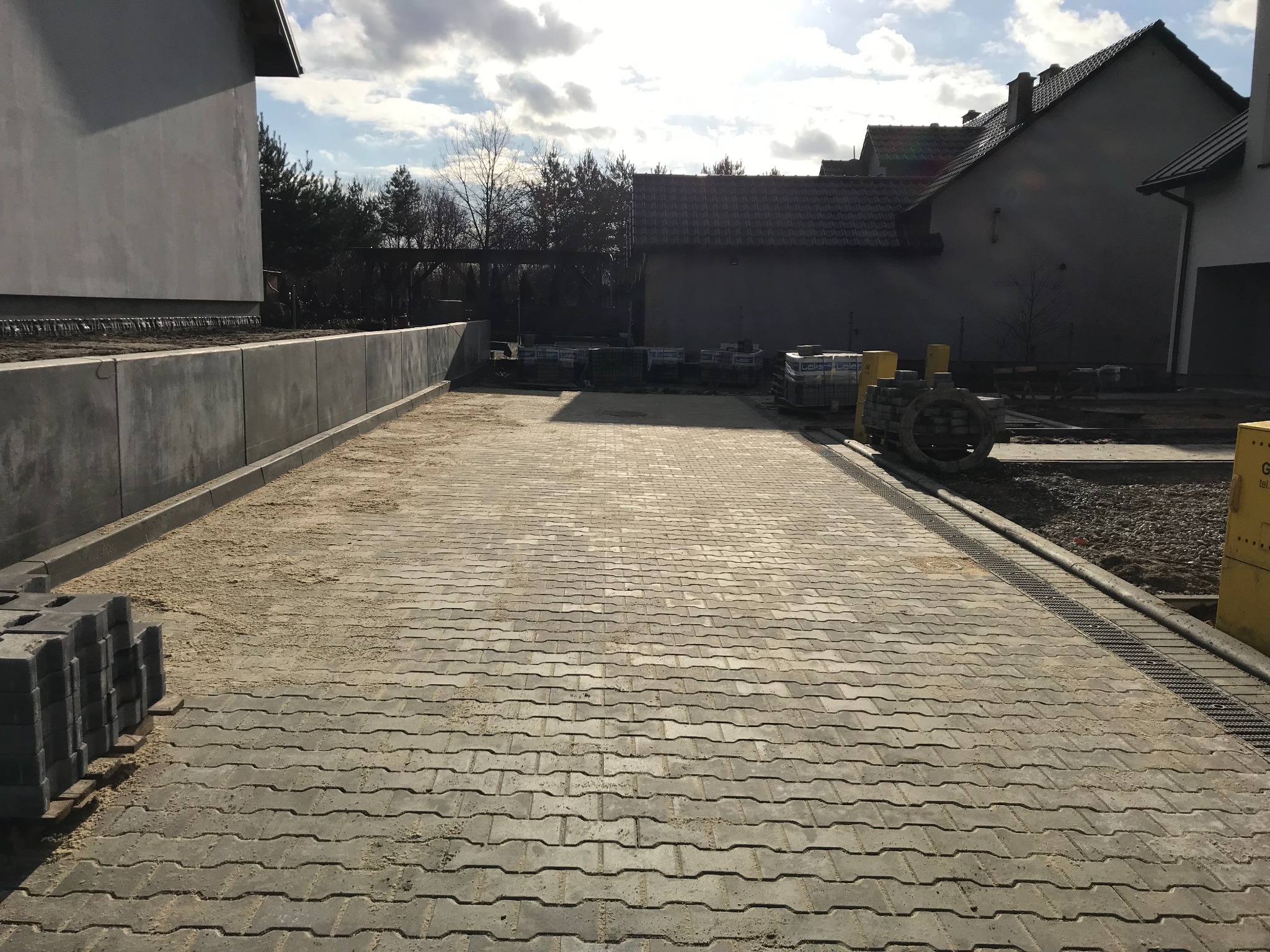 Ułożona szara kostka brukowa na podjeździe, widoczne palety z kostką w tle, betonowe obrzeża i odwodnienie liniowe. Częściowo zasypana piaskiem.
