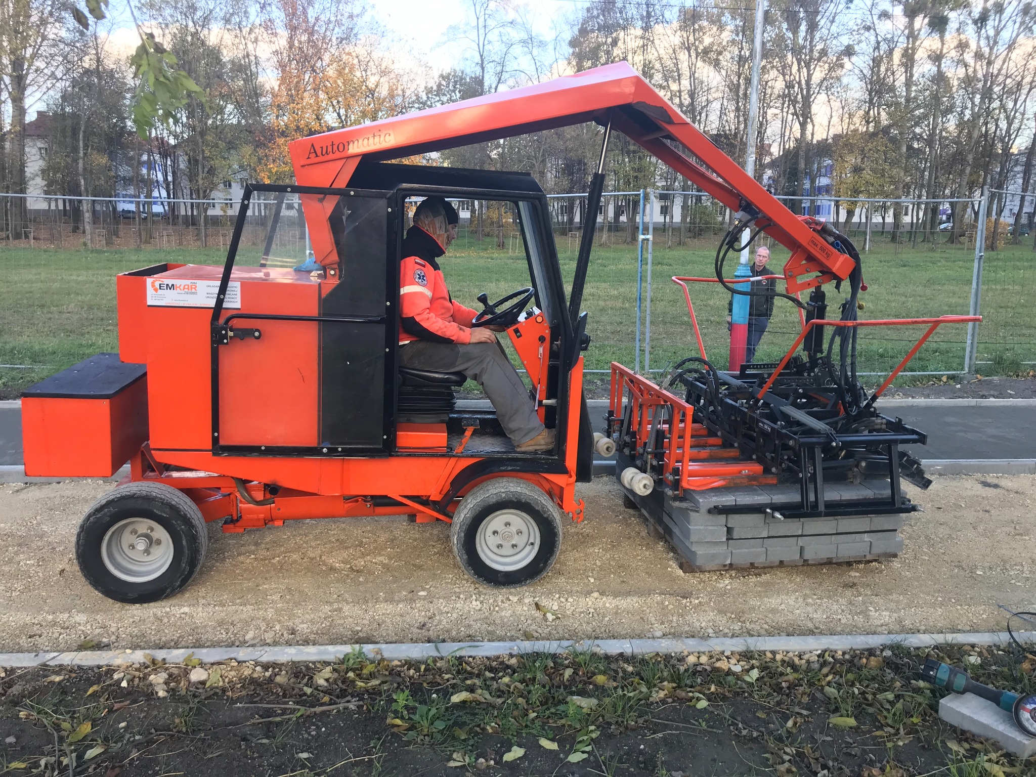 Maszyna do układania kostki brukowej w akcji: operator w kabinie, kostka podawana automatycznie. Widoczne narzędzia i osoby w tle. Praca w toku.