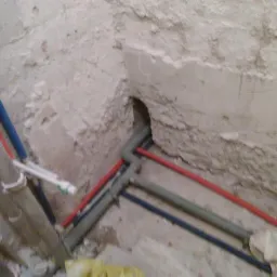 Surowe ściany z widocznymi rurami wodnymi w kolorze czerwonym, niebieskim i szarym, wychodzącymi z otworu w ścianie, w trakcie montażu instalacji.