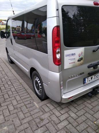 Srebrny bus marki Opel Vivaro zaparkowany na chodniku z kostki, z naklejką reklamową firmy przewozowej Ganderbus na tylnych drzwiach.