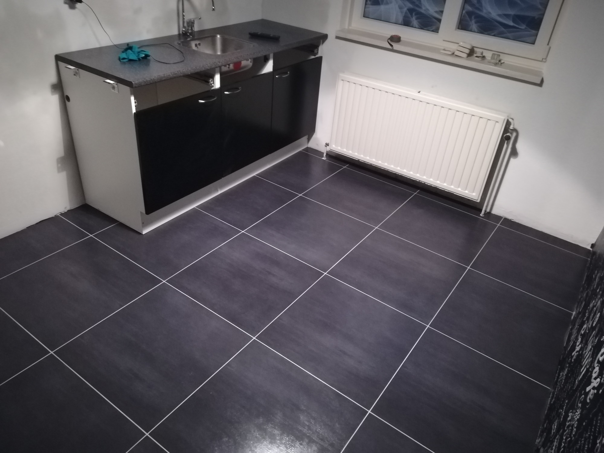 Ukończona łazienka z ciemnymi, kwadratowymi płytkami podłogowymi z jasnymi fugami, biały grzejnik pod oknem i szafka podumywalkowa z czarnymi frontami i szarym blatem.