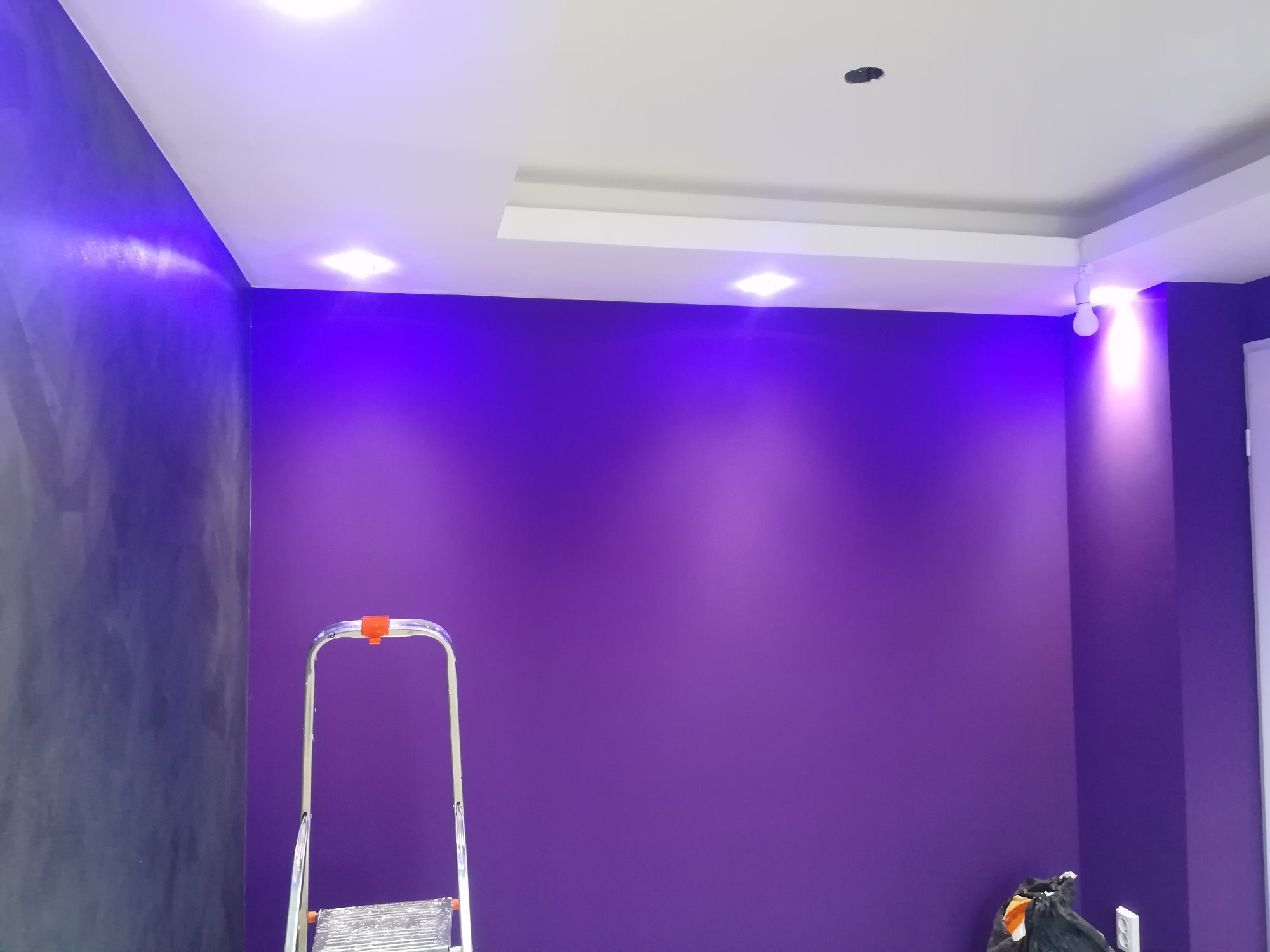 Pomieszczenie po malowaniu ścian na fioletowo, widoczna drabina i wiadro z farbą, sufit z oświetleniem punktowym i elementami dekoracyjnymi.