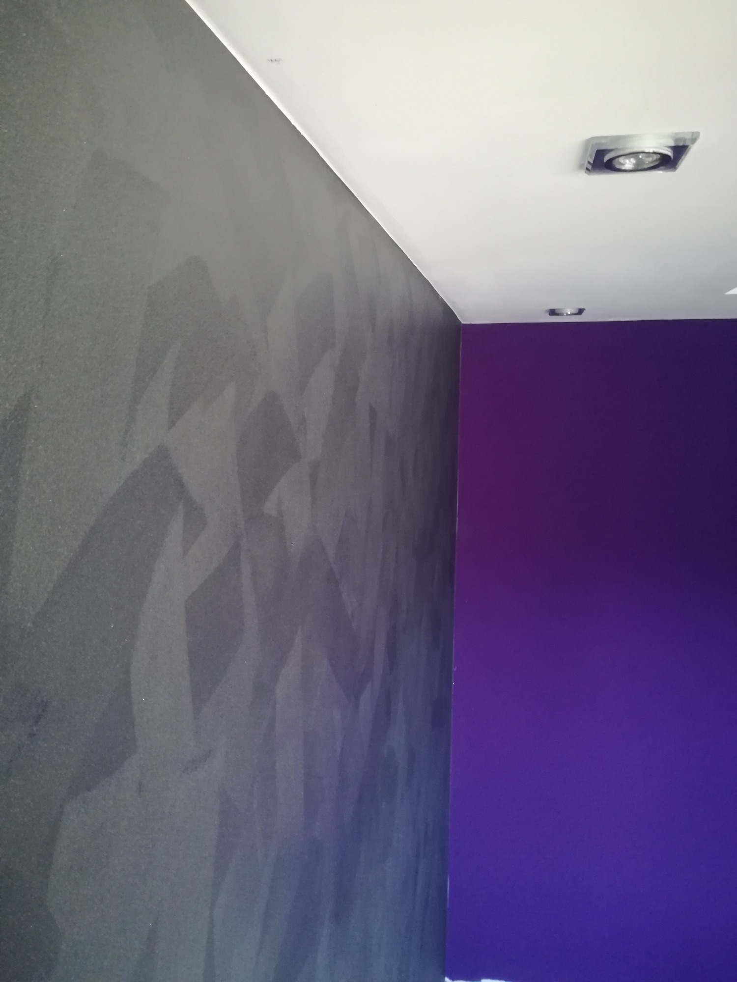Ściana z dekoracyjną szarą strukturą przypominającą beton architektoniczny sąsiadująca z gładką, fioletową ścianą, oświetlona punktowym światłem.