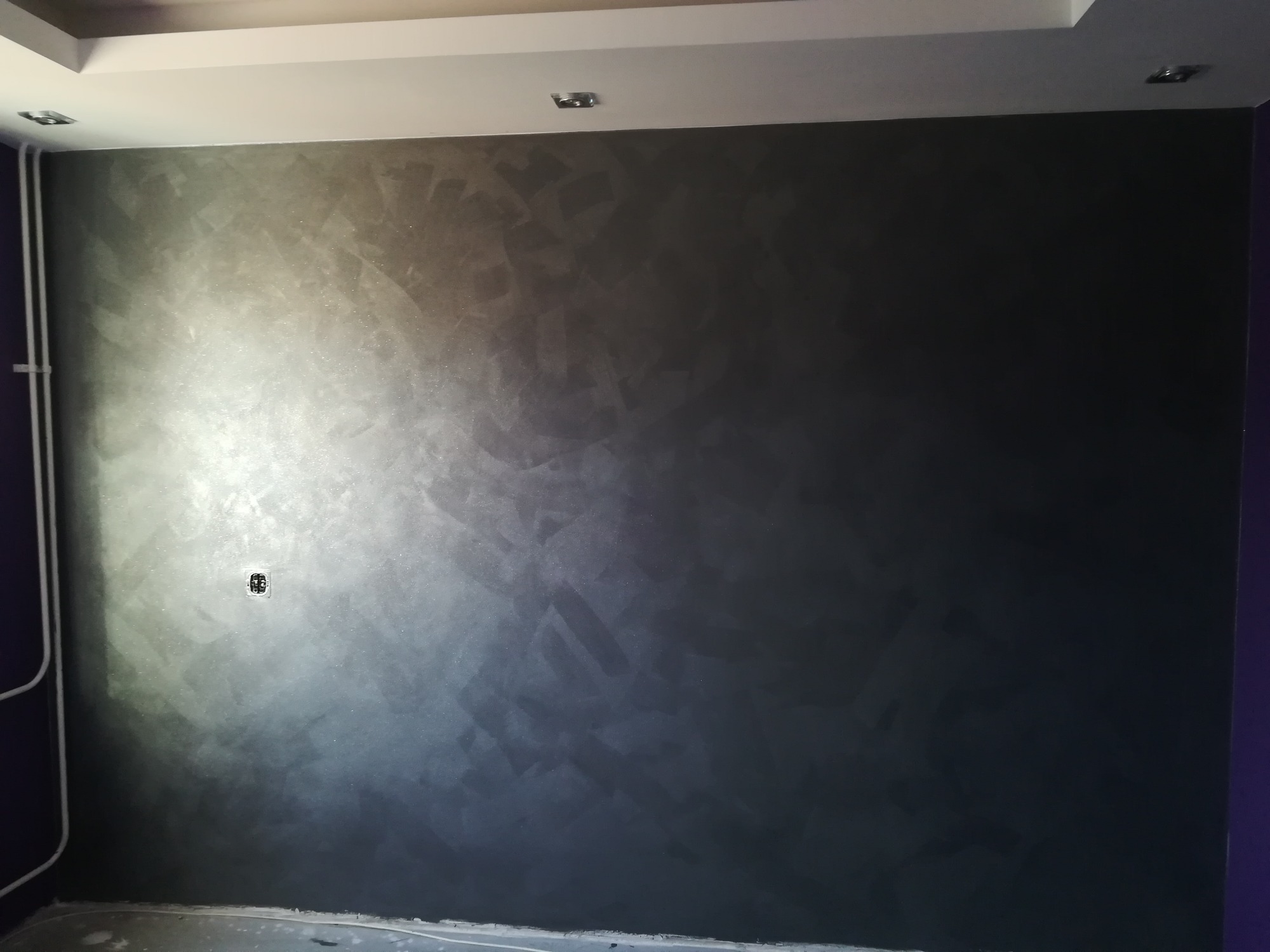 Ściana w odcieniach szarości z dekoracyjną fakturą, gładź w stylu weneckim, widoczne gniazdko elektryczne i fragment instalacji przy ścianie w kolorze fioletowym.