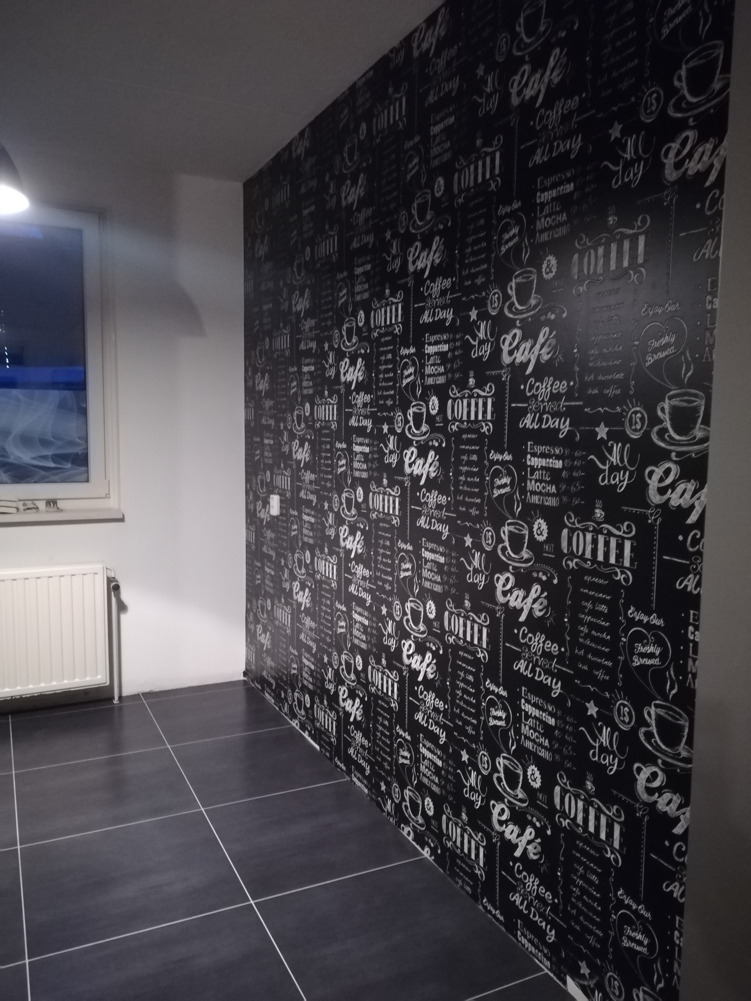 Ściana wykończona tapetą z motywem kawowym w czarno-białej kolorystyce, wzór zawiera napisy i rysunki filiżanek, na podłodze ciemne płytki, widoczny fragment grzejnika i okna.