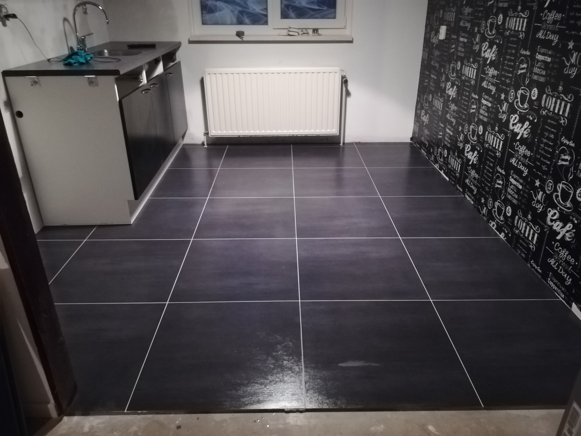 Wykończona kuchnia z czarną podłogą z płytek, białą zabudową kuchenną, grzejnikiem pod oknem i ścianą z tapetą z motywami kawowymi.