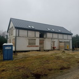 DNM ANNA SPYSZEWSKA - Dwukondygnacyjny budynek w stanie surowym zamkniętym z szarej cegły, z ciemnoszarym dachem z oknami dachowymi, zabezpieczone deskami otwory okienne, na zewnątrz przenośna toaleta.