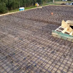 Siatka zbrojeniowa fundamentu na deskowaniu z widocznymi plastikowymi dystansami, przygotowana do zalania betonem, w tle teren budowy z drzewami i małą architekturą.