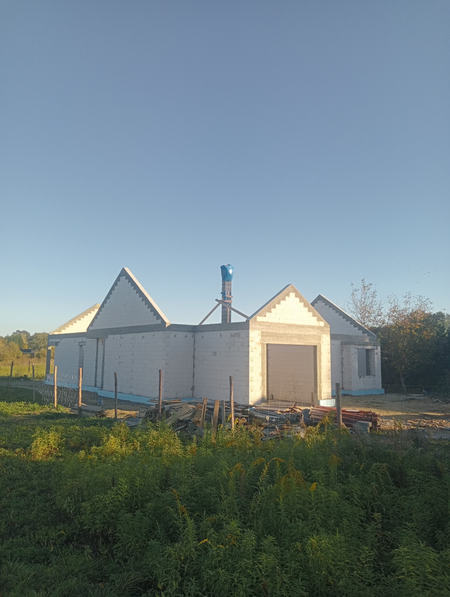 Niedokończony dom murowany z białych bloczków, widoczna izolacja fundamentów i częściowo wykonane nadproża okien i bramy garażowej, na dachu komin zabezpieczony folią.